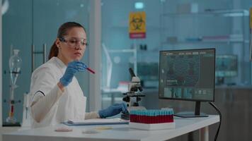 Scientific researcher looking at substance in test tube and working on computer for development in laboratory. Woman biologist analyzing red liquid in glass flask, using monitor in lab. video