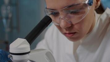 Woman looking through microscope lens to analyze dna sample in laboratory. Scientist using microscopic tool with magnifying glass to examine substance and genetic. Handheld shot. Close up video
