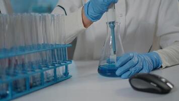 Biologist using micro pipette to mix substance in beaker and test tube, working with lab glassware in laboratory. Woman scientist holding dropper for science experiment. Close up video