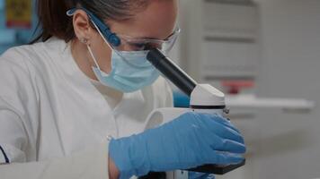 Biologist with face mask using microscope in science laboratory, analyzing dna sample and substance with microscopic tool. Woman with protective glasses working with magnifying glass lens video