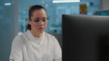 Woman chemist working on computer in science laboratory, using technology to develop microbiology experiment with lab equipment. Scientist with protective glasses. Handheld shot video