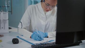 Woman chemist taking notes and using computer for experiment in laboratory. Specialist working on scientific development, doing research study and writing on paper in science lab. video