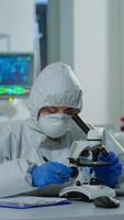 Vertical . Medical research scientist conducting DNA experiments under microscope wearing protection suit and writing informations on clipboard. Chemist using high tech for vaccine development against covid19 video