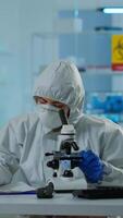 Vertical . Biologist in ppe suit working on microscope and writing information on clipboard in equipped laboratory. Chemist examining vaccine evolution using high tech researching diagnosis against covid19 virus video