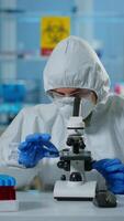 Vertical . Scientist in ppe suit making adjustments and looking through laboratory microscope, typing on pc. Chemist in coverall working with various bacteria, tissue blood samples for antibiotics research video