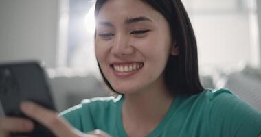 Footage close-up of Happy young Asian woman smiling and using smartphone on big comfy sofa at living room. People, technology and lifestyle concepts. video