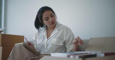 Handheld shot, Asian young woman sits and checking books in box and write on notebook after move into new home video