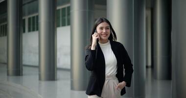 Front view shot, Footage of Young smiling elegant Asian busy business woman in a suit talking with a smartphone and walking in front of a modern business building. Business and people concepts. video