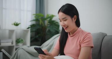 Footage of Happy young asian woman relax on comfortable couch at home shopping online on smartphone, smiling girl use cell phone chatting, browse wireless internet on gadget video