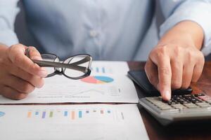 Close-up of hand holding a glasses reviewing the chart, graph report and calculating on the calculator for planning next step of the business. Space for text. Business, finance, and planning concept photo