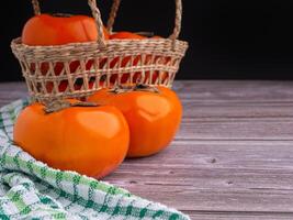 Fresh ripe persimmons in a basket placed on a wooden table with a black wall background. Space for text. Close-up photo. Concept of healthy fruit photo