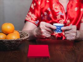 Hands of a senior woman wearing a traditional cheongsam qipao dress holding a teacup with red envelopes or Ang Pao and four tangerines in a basket placed on a wooden table. photo
