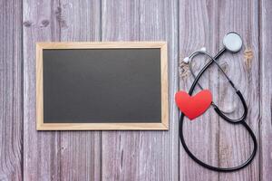 Top view of a small blackboard with a stethoscope and red heart paper placed on a wooden table. Space for text. Concept of love and healthcare photo