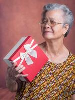 Portrait of an elderly Asian woman holding a red gift box and looking at the camera while standing in the studio. Concept of the festival, anniversary and aged people photo