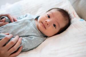 un adorable asiático recién nacido bebé hijo en azul pijama es en su cama, hablando y jugando con su mamá. foto