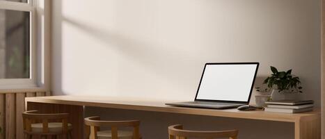 A laptop computer with a white-screen mockup on a long wooden table in a comfortable minimalist cafe photo