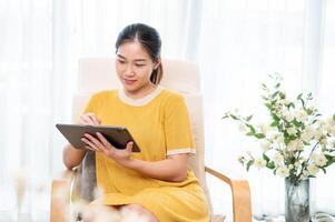 un hermosa asiático mujer utilizando su digital tableta en un Sillón en el vivo habitación. foto