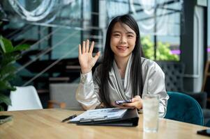 A confident Asian businesswoman is waving her hand and smiling at the camera while sitting at a desk photo