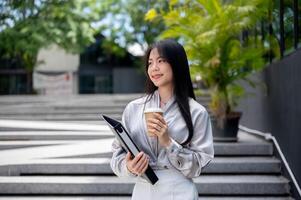 un contento asiático mujer de negocios con un archivo carpeta y un para llevar café taza es caminando abajo el escaleras. foto