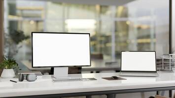 A computer desk in a modern office room features a computer and a laptop with white-screen mockup. photo