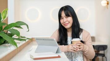 un atractivo asiático mujer es sonriente a el cámara, sentado a un mesa en un café con su tableta. foto