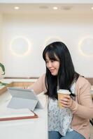 un atractivo asiático mujer teniendo un caliente bebida y utilizando su digital tableta en un minimalista cafetería. foto