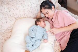un feliz, cuidando joven asiático mamá es hablando, jugando, y consolador su recién nacido bebé en el cama. foto