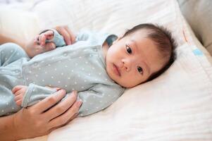 un adorable asiático recién nacido bebé hijo en azul pijama es en su cama, hablando y jugando con su mamá. foto