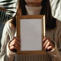 Woman in a cozy sweater holding a blank picture frame, ready for customization. Warm lighting and soft textures create a welcoming atmosphere. photo
