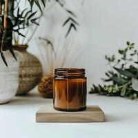 Minimalist amber candle on wooden block, with dried plants and green foliage in background, set on white surface. photo