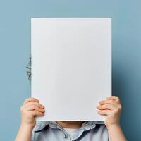 Child holding blank white sheet of paper hiding face standing against blue background. Creativity, innocence, copy space concept. photo