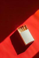 Minimalistic image of a white matchbox with orange-tipped matches on a red background, bathed in bright sunlight casting shadows. photo