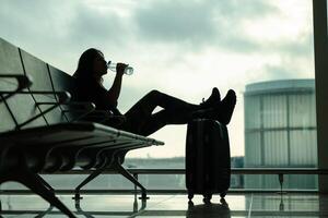 Woman sitting on an airport bench, drinking a bottle of water while waiting for her flight, with a background of sky with clouds and low angle shot photo