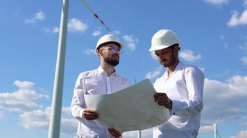 två multietnisk ingenjörer diskuterar en plan på en vind bruka i främre av en turbin levererar alternativ elektricitet från de omvandling av kinetisk energi video