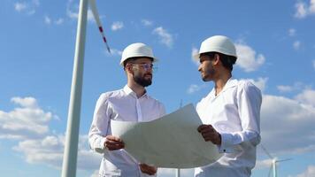 två multietnisk ingenjörer diskuterar en plan på en vind bruka i främre av en turbin levererar alternativ elektricitet från de omvandling av kinetisk energi video