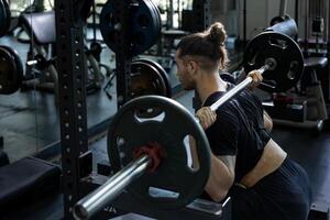 Caucasian muscular body builder sport man is practice weight training on squat barbell for core muscle inside gym with dark background for exercising and workout photo