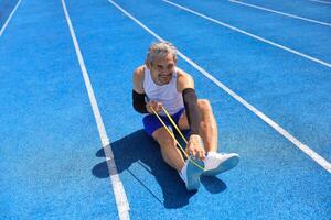 Senior Asian athlete using stretching rubber band warm up for running in the professional blue track for professional exercise, sport training workout and healthy for longevity photo