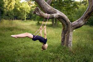 a little 5-year-old gymnast in a black minimalist jumpsuit swings on a gymnastic rope on a tree photo
