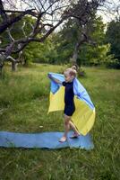 a little 5-year-old gymnast in a black minimalist jumpsuit is practicing with the flag of Ukraine in the garden photo