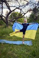 a little 5-year-old gymnast in a black minimalist jumpsuit is practicing with the flag of Ukraine in the garden photo