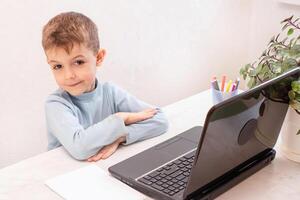 close-up of a happy boy in a blue sweater looking into the frame while sitting near a laptop and waiting for the online lesson to start. teaching people at home in bedroom, studying online on computer photo
