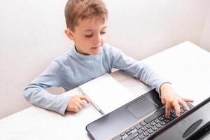 child student returns to school. boy smiles, writes or draws and reads on laptop while doing homework. teaching people at home in bedroom, studying online on computer photo