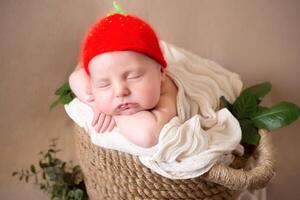 a cute newborn iin a hat of strawberries in the form of a sunflower flower sleeps peacefully. beige background with space for text with a baby sleeping in a wicker basket decorated with green leaves. photo