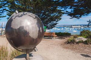 Metallic globe sculpture on stand with reflective surface featuring continents outlined against clear blue sky. Harbor view with boats and lush greenery in background. Peaceful coastal setting. photo