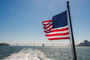 American flag on pole flutters against blue sky, Golden Gate Bridge visible in background over San Francisco Bay. City skyline faintly visible to left. Prideful composition. photo