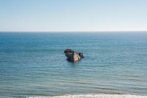 Serene seascape under clear blue skies with calm ocean waters and a shipwreck visible. Rusted and weathered remains suggest age. Tranquil and remote coastline scene. photo
