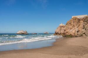 Tranquil San Francisco beach scene with sandy shore, lapping waves, rocks, and a cliff with a building on top. Peaceful ambiance with few individuals. photo