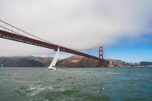Serene maritime scene in San Francisco with the Golden Gate Bridge in the backdrop. White sailboat, partly cloudy sky, and misty hills add natural mystique. photo