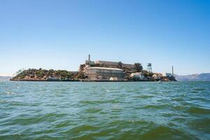 Alcatraz Island view under clear sunny skies, featuring Alcatraz Federal Penitentiary structures from the bay in San Francisco Bay. Notable landmarks visible. photo