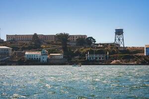 Clear day view of Alcatraz Island in San Francisco Bay. Image shows main prison building, other structures, rugged terrain, blue waters, and water tower. photo